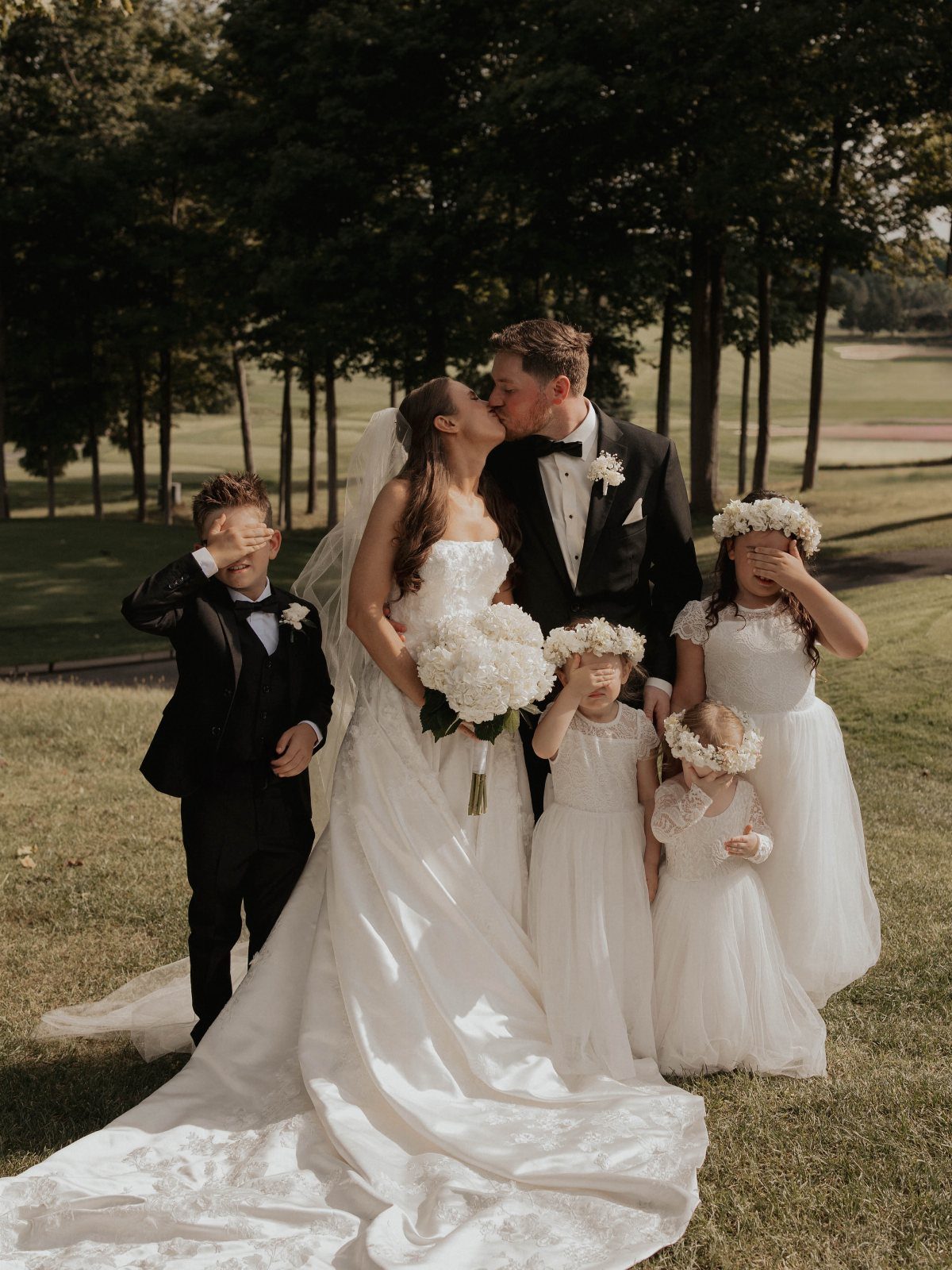 Bride and groom kissing while children in wedding attire cover their eyes.