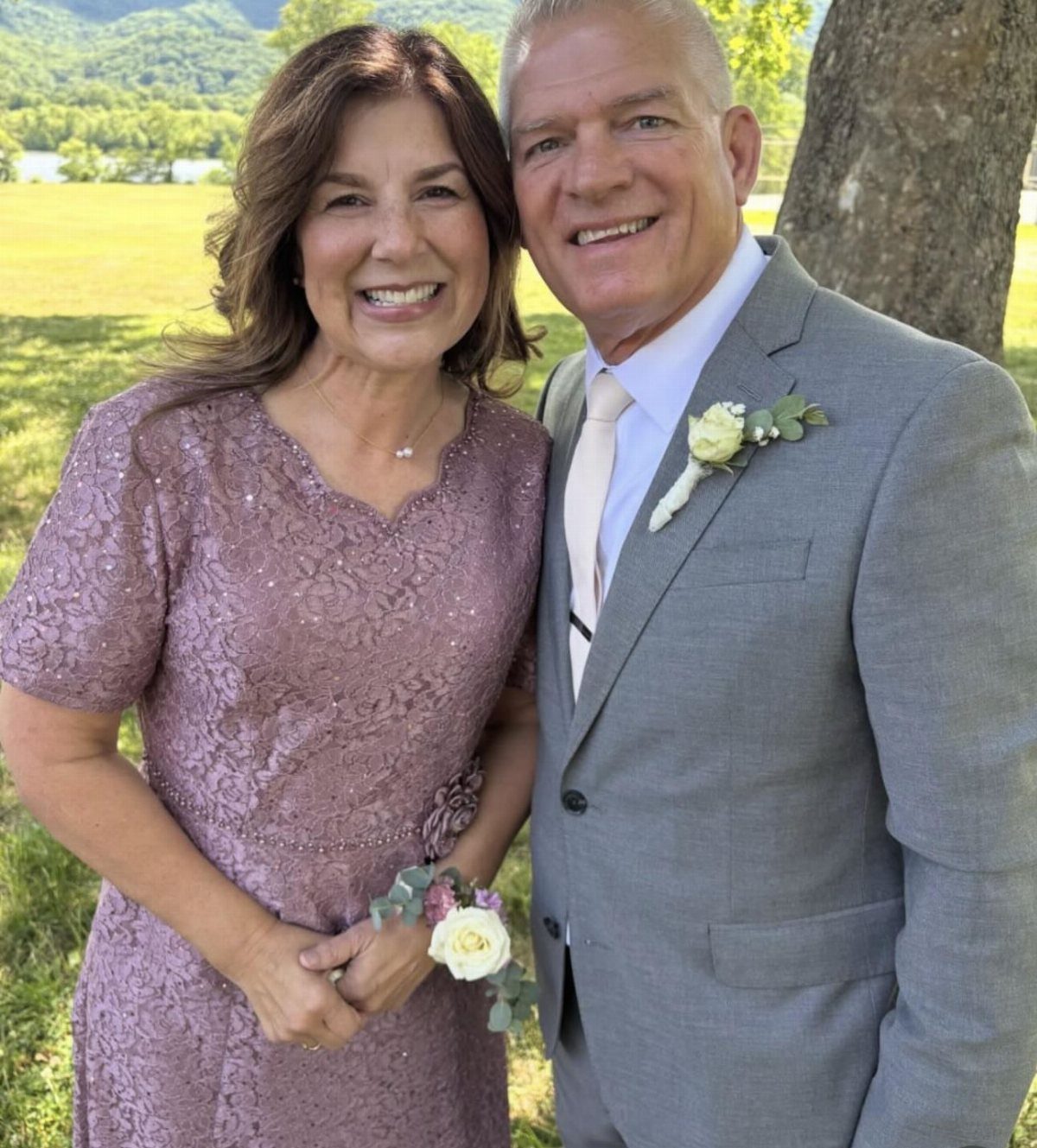 Smiling couple in formal wedding attire posing outdoors against a green background.