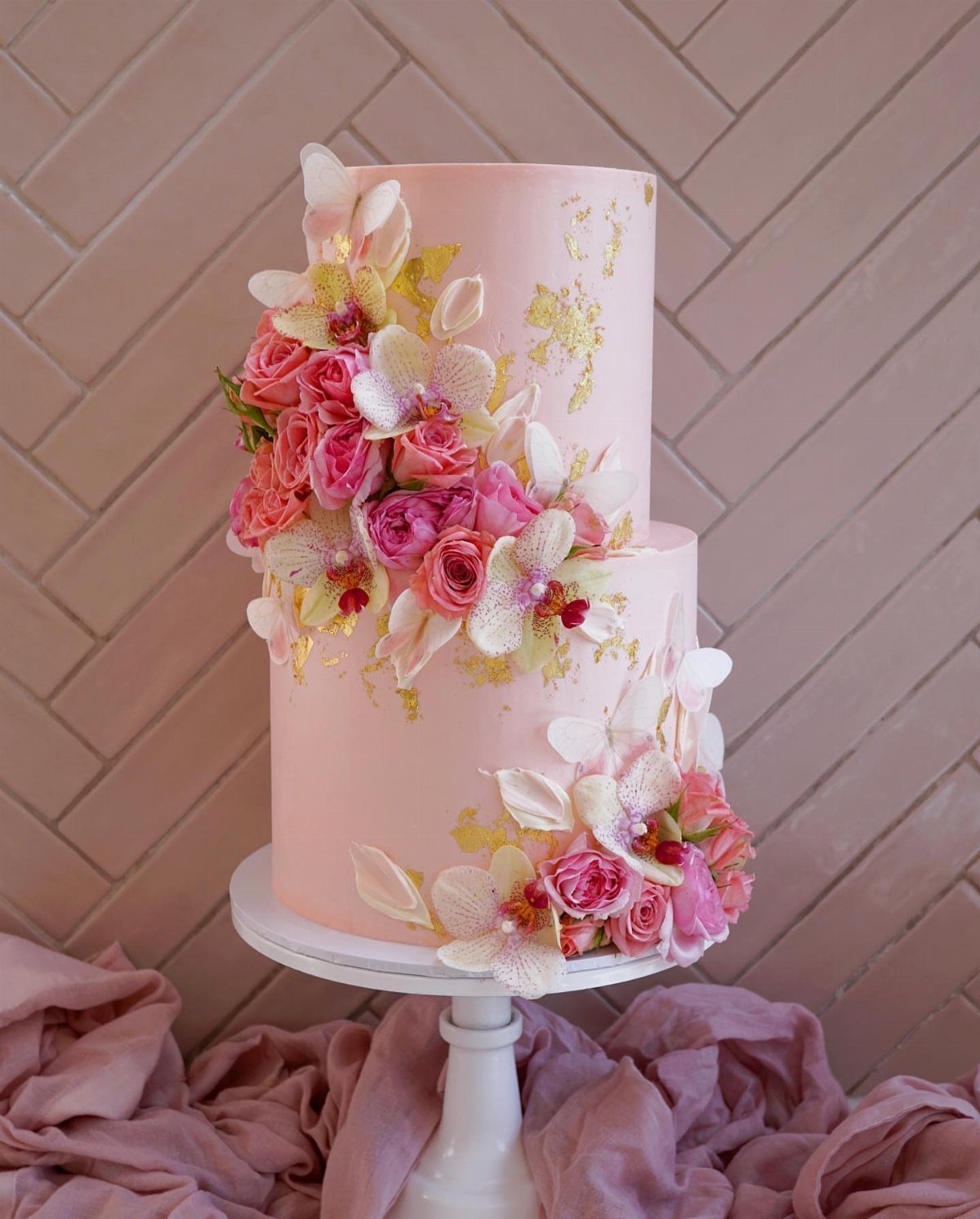 Two-tier pink cake decorated with pink roses, white orchids, and gold leaf accents.