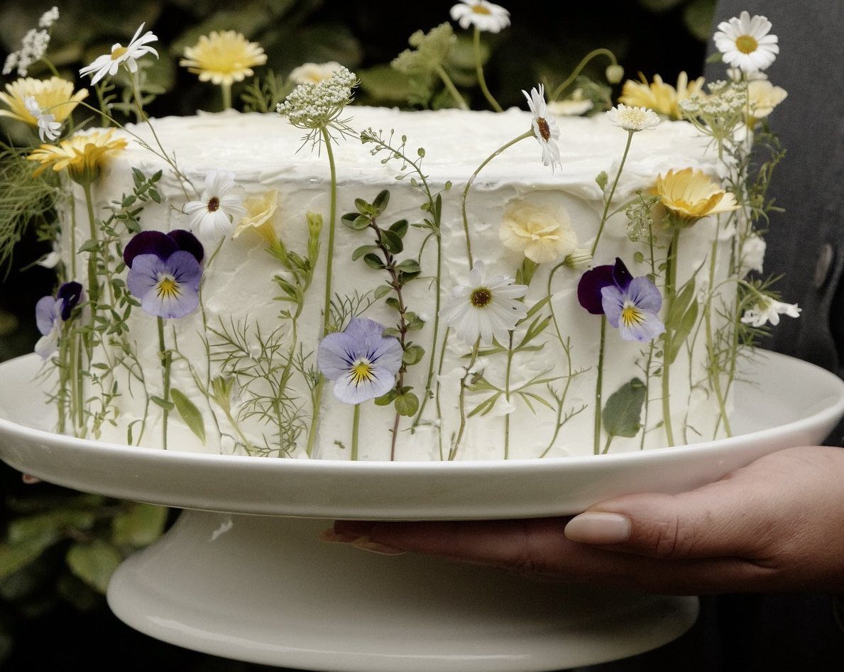 White frosted cake decorated with pressed wildflowers and greenery on a white stand