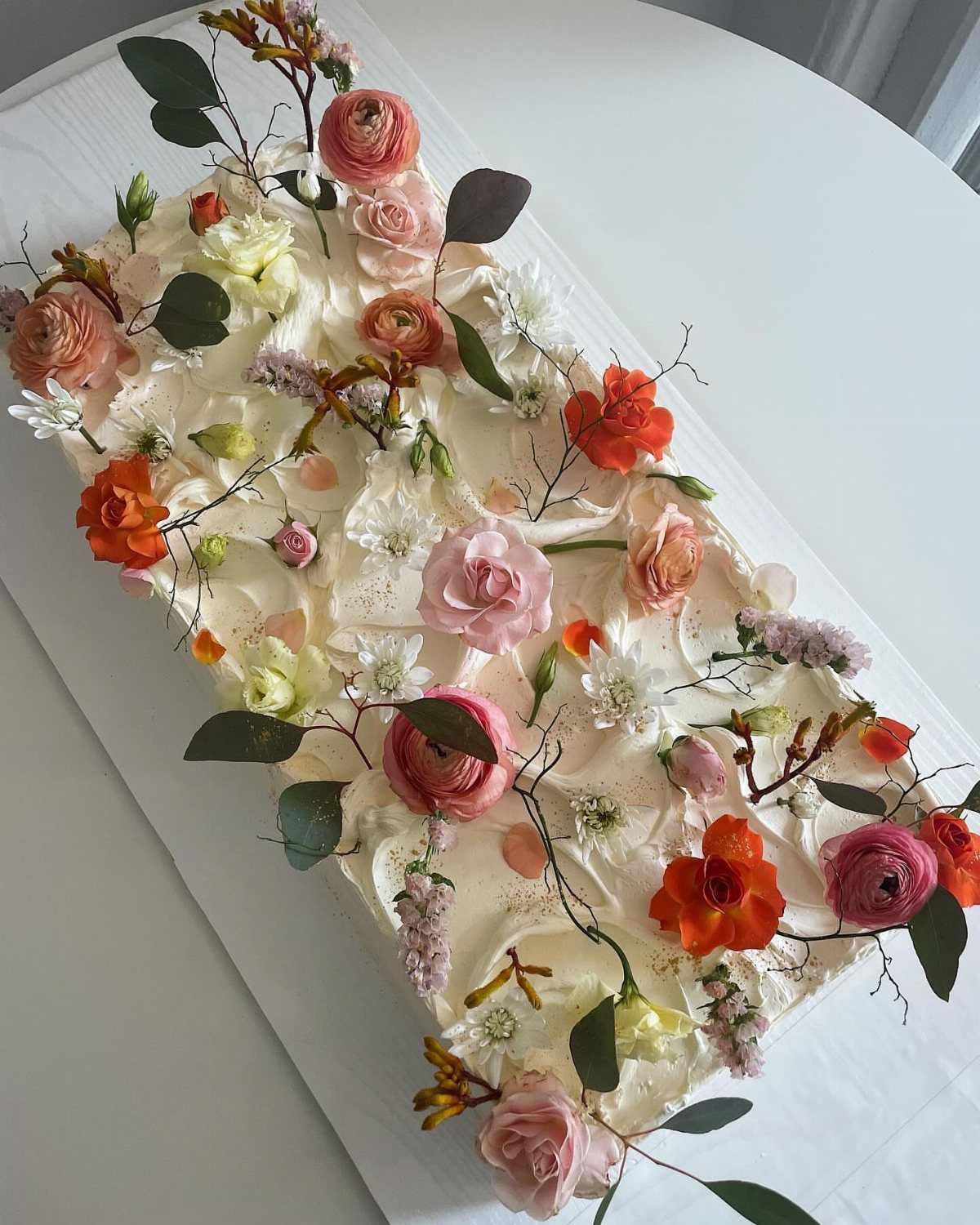 White frosted rectangular cake decorated with scattered pink and orange fresh flowers and greenery.