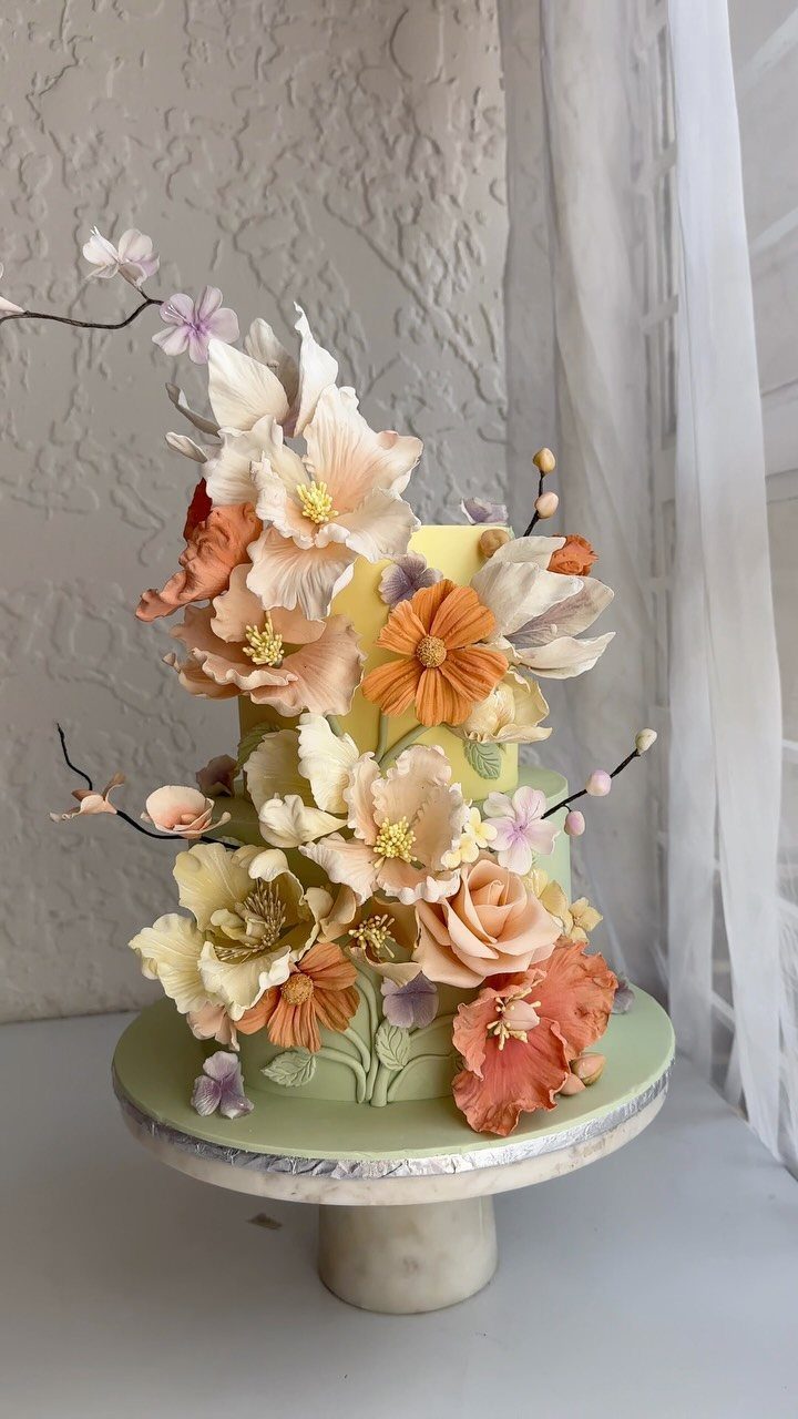 Multi-tiered yellow and green cake adorned with realistic peach and orange sugar flowers.