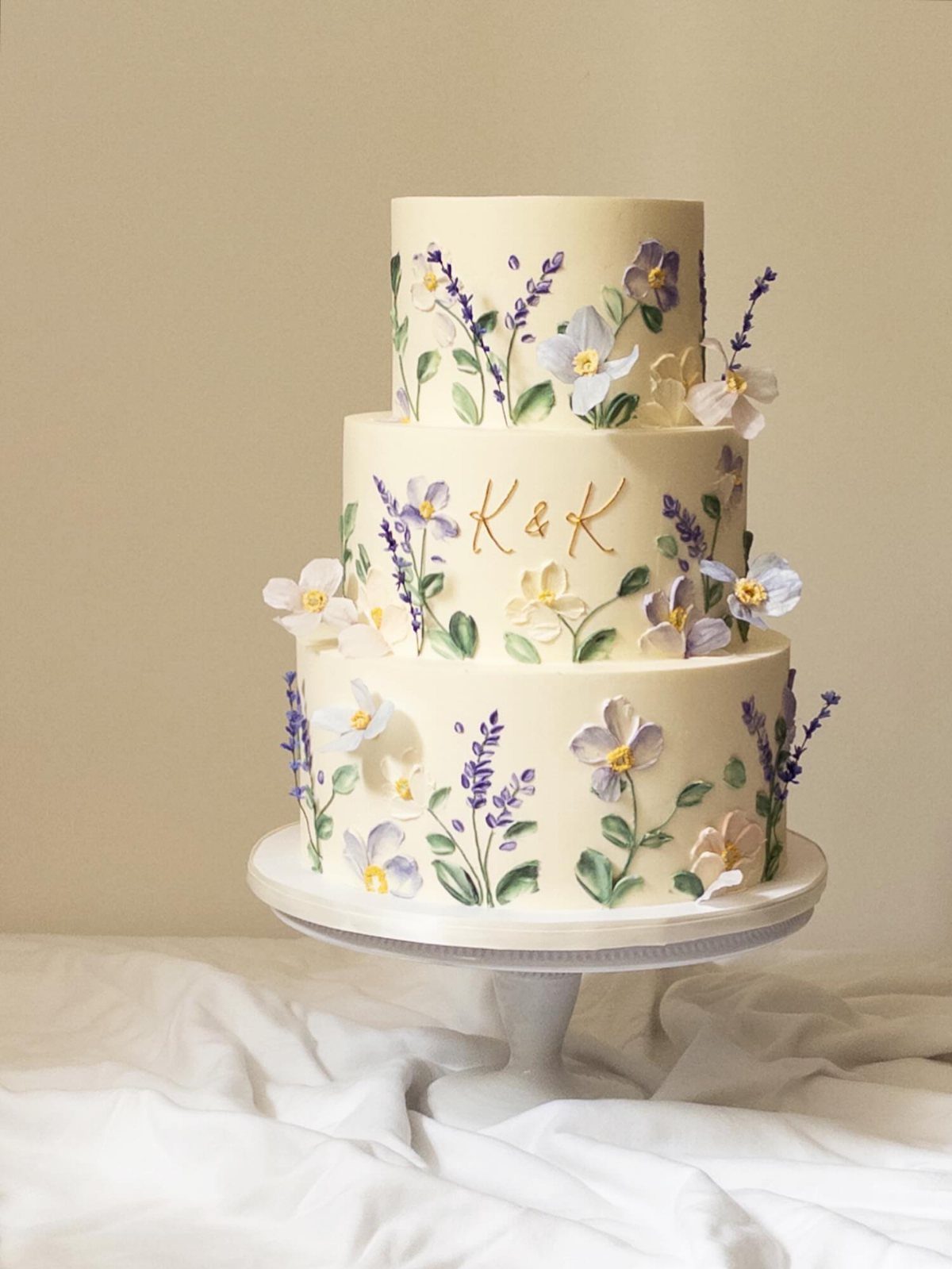 Three-tiered white wedding cake decorated with purple and white flowers and gold initials.