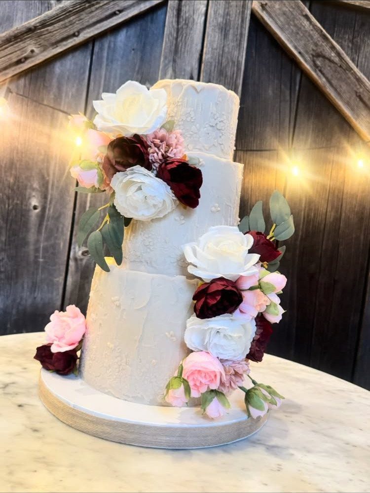 Three-tiered white wedding cake with burgundy and pink roses against a rustic barn background