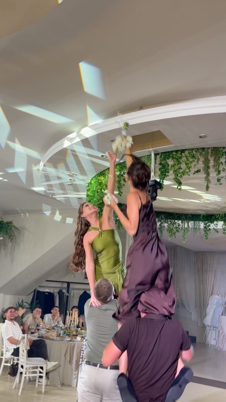 Two women lifted on shoulders reaching for a wedding bouquet hanging from the ceiling.