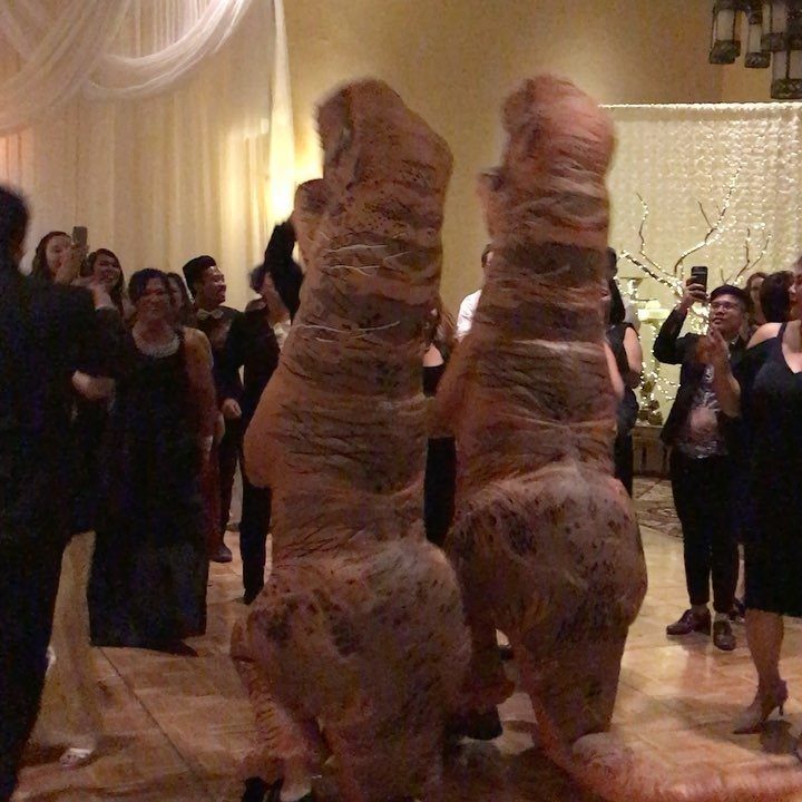 Two people in dinosaur costumes standing in a ballroom surrounded by formal guests.