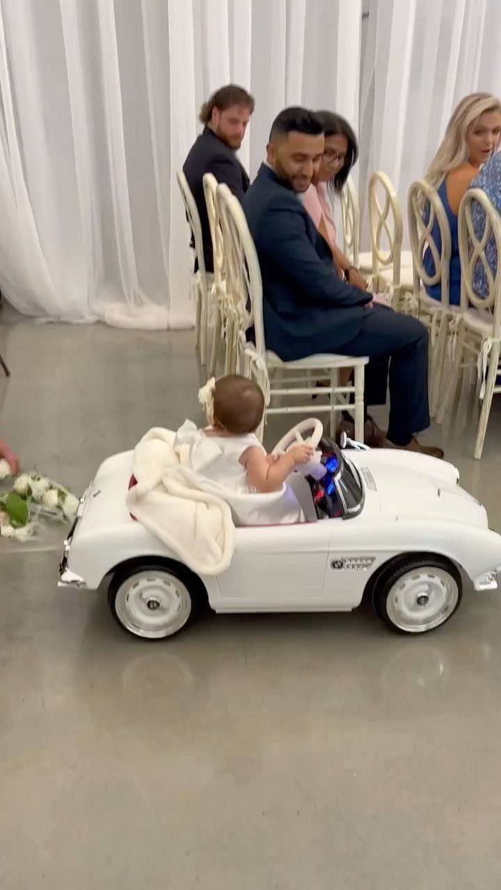 Small child in white dress sitting in a white toy convertible car at wedding