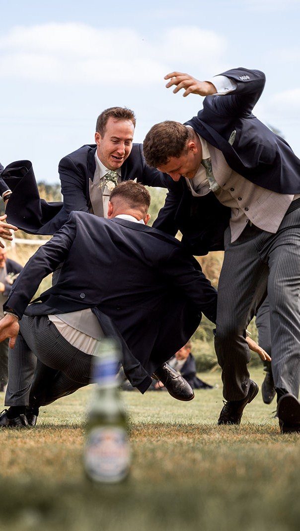 Groomsmen in navy suits tumbling and falling over each other on a grassy field.