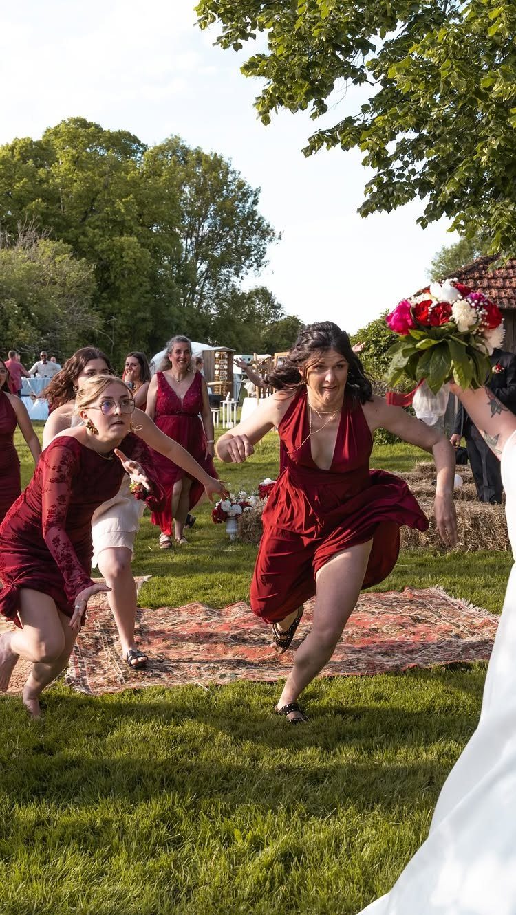 Bridesmaids in red dresses running and reaching for a wedding bouquet outdoors.