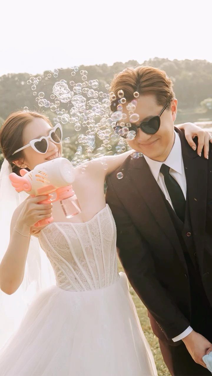 Bride in heart sunglasses blowing bubbles toward groom in a black suit outdoors.