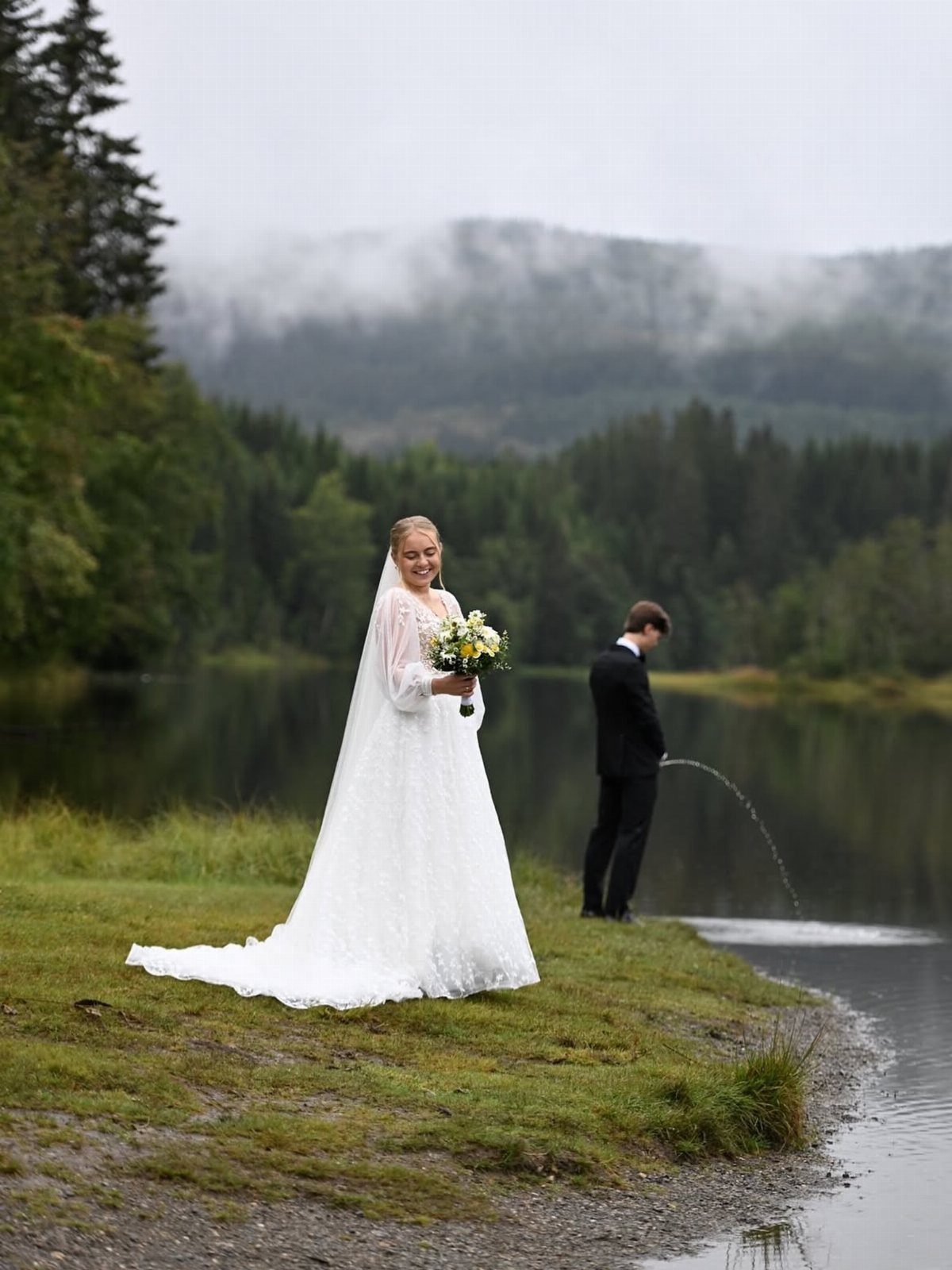 Bride smiling in white dress while groom pees into a lake nearby.