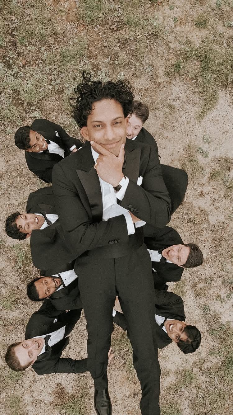 Groom in black tuxedo posing while groomsmen peek out from behind him outdoors.