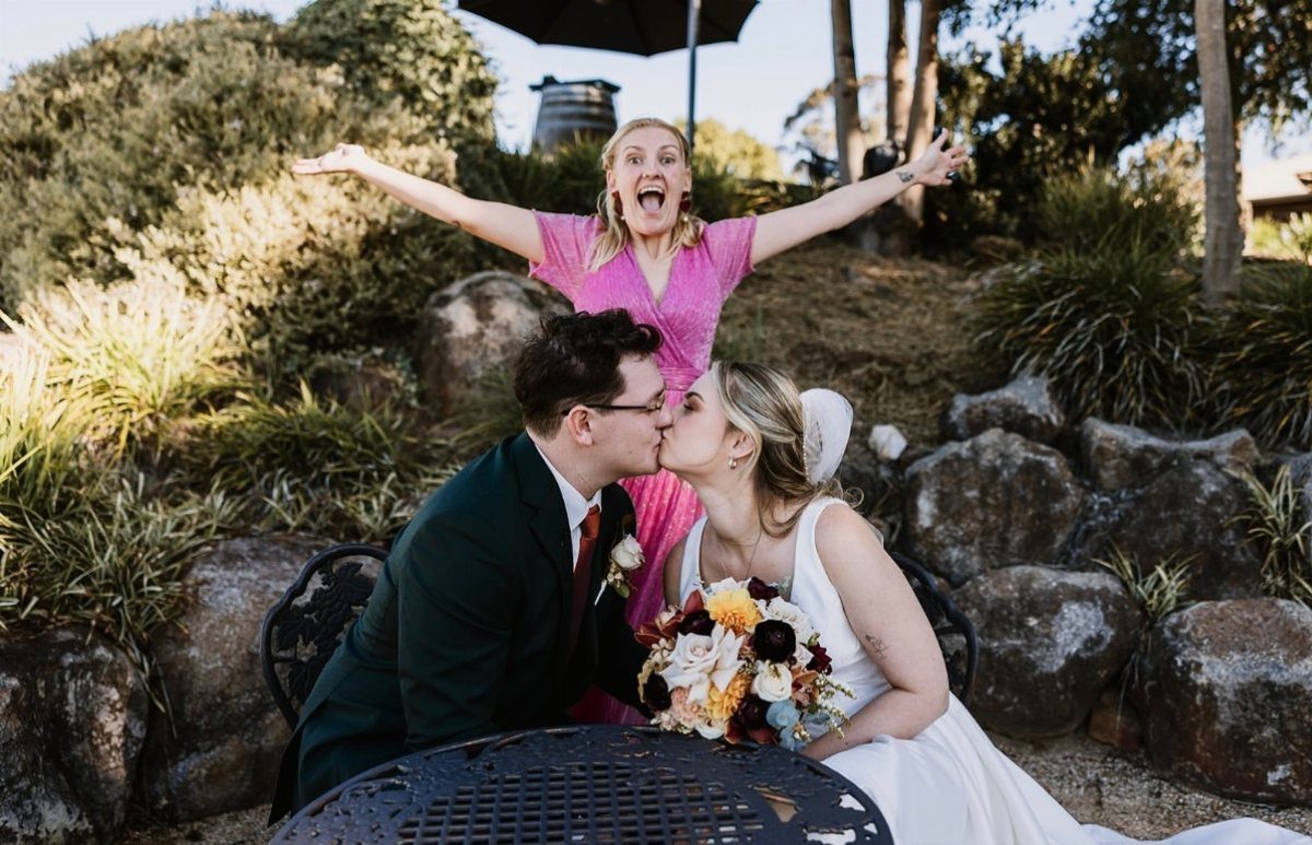 Bride and groom kissing while a woman in pink makes a shocked face behind them.