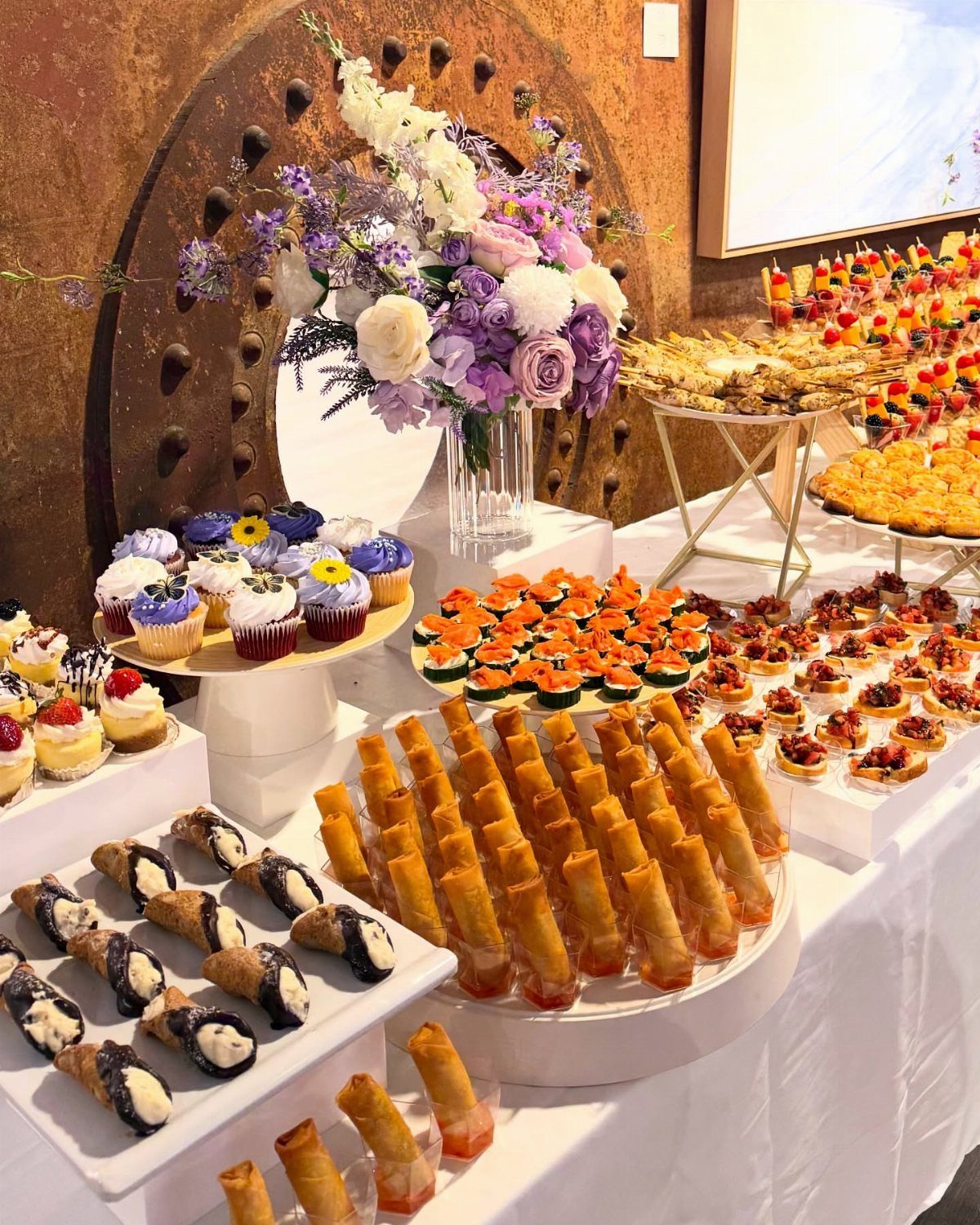 Food spread with cupcakes, savory appetizers, and spring rolls next to purple flowers.