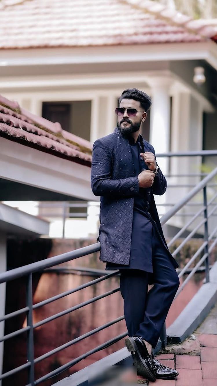 Man in dark blue sherwani and sunglasses posing by a metal railing outdoors.