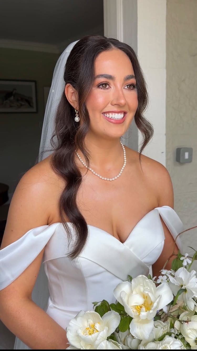 Bride with natural makeup, wearing a white dress and pearls, holding white flowers.