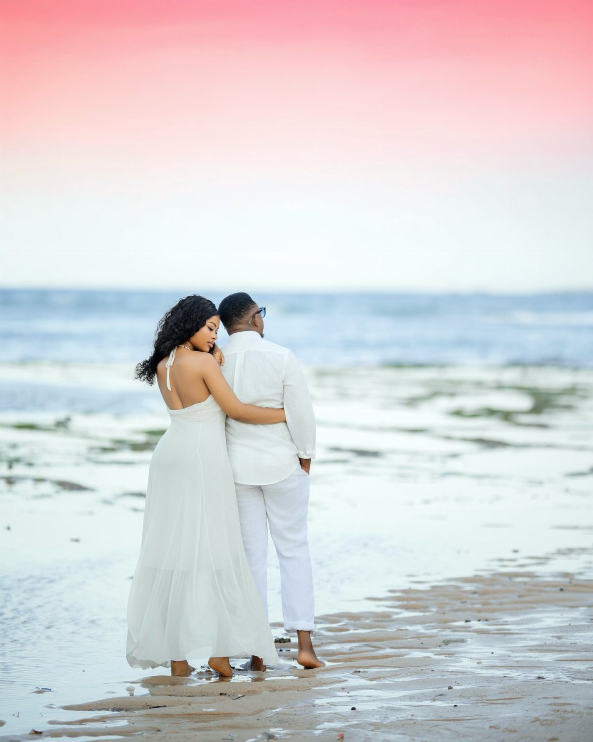 Couple in white outfits hugging on a beach under a pink sunset sky.