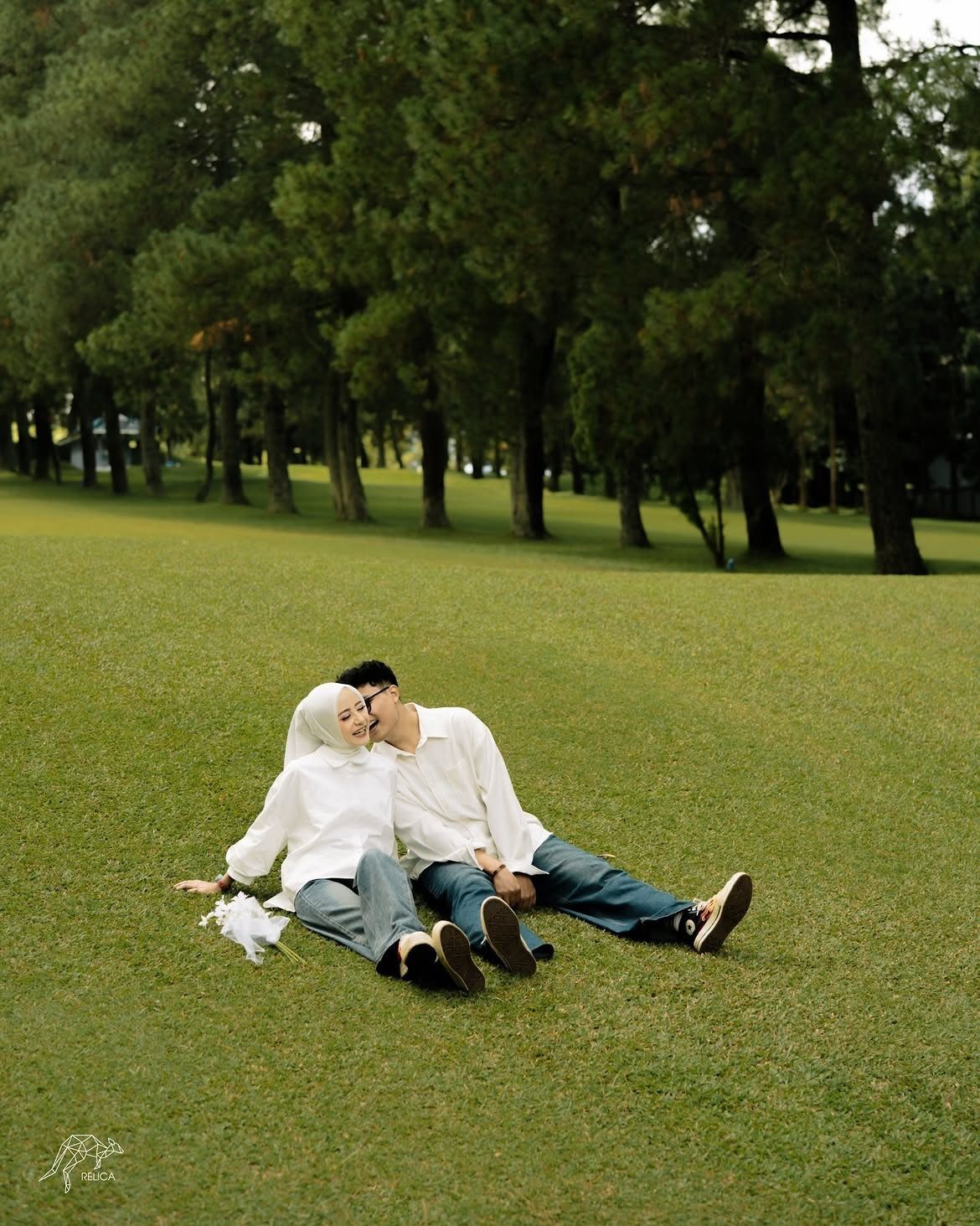 Couple in white shirts and jeans sitting on grass in a wooded park
