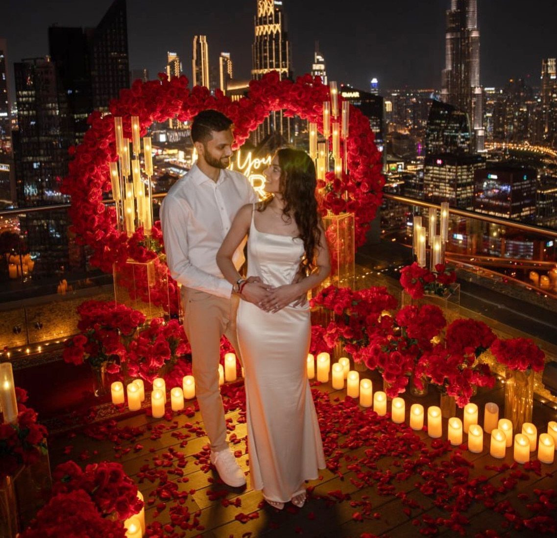 Couple posing on a rooftop with red roses and candles against a city skyline.