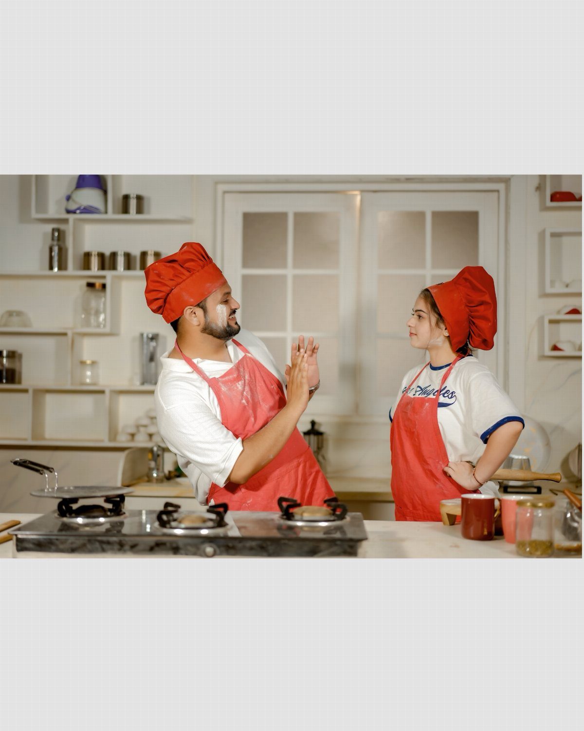 Couple in red chef hats and aprons smiling in a white kitchen