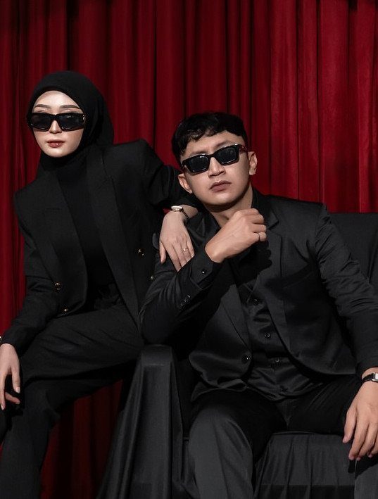 Couple in black suits and sunglasses posing against a red curtain backdrop.