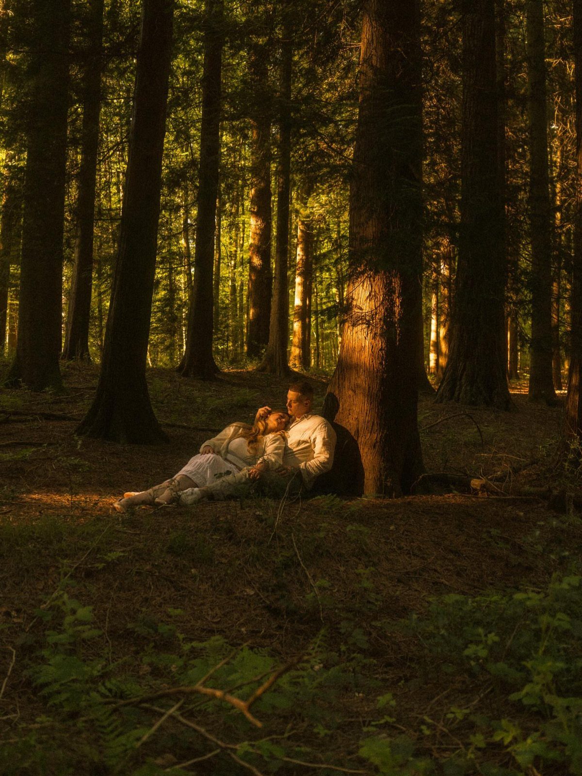 Couple reclining against a large tree in a sun-drenched, dense forest setting.