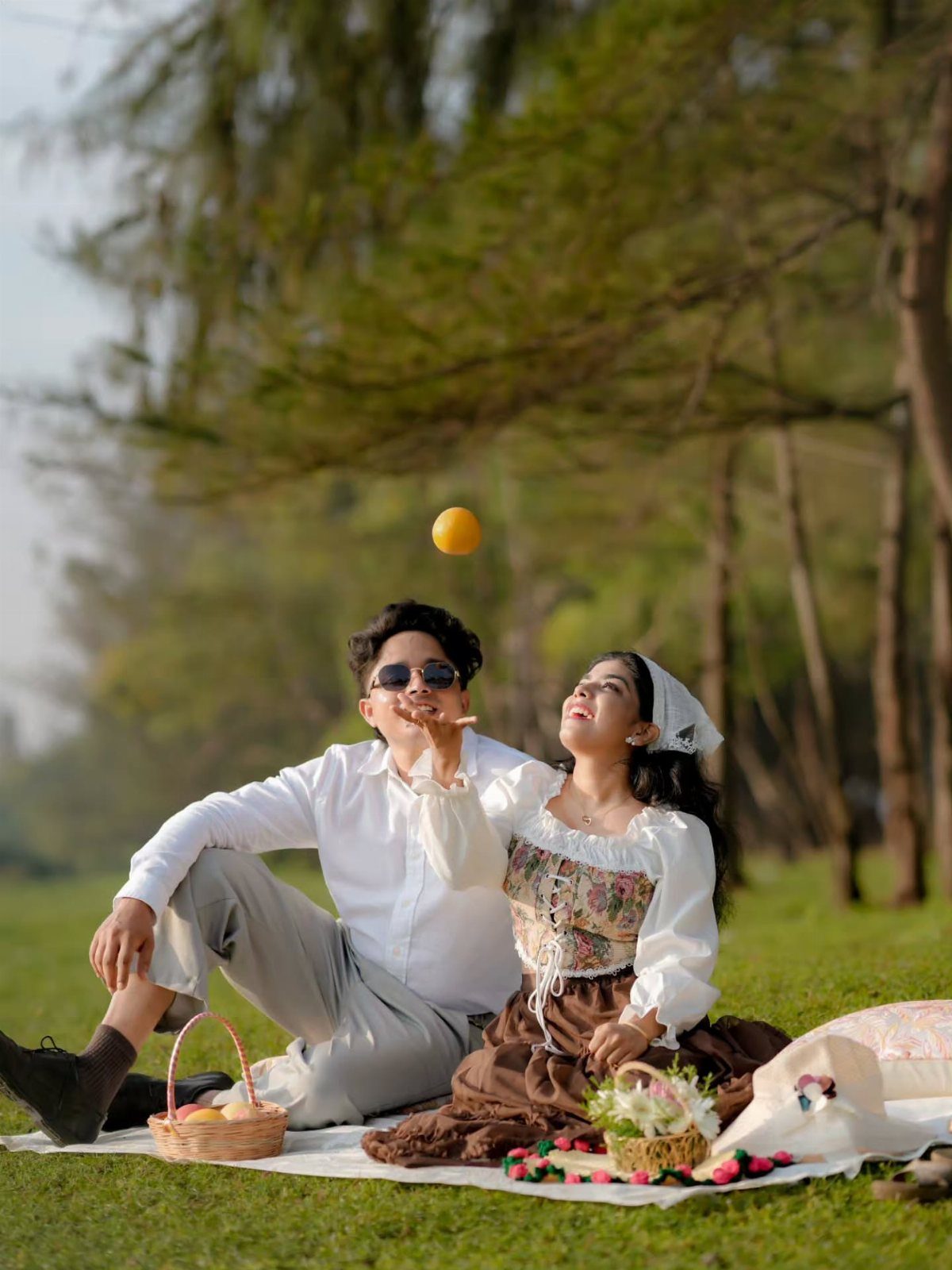 Couple sitting on a picnic blanket in a park tossing an orange together.