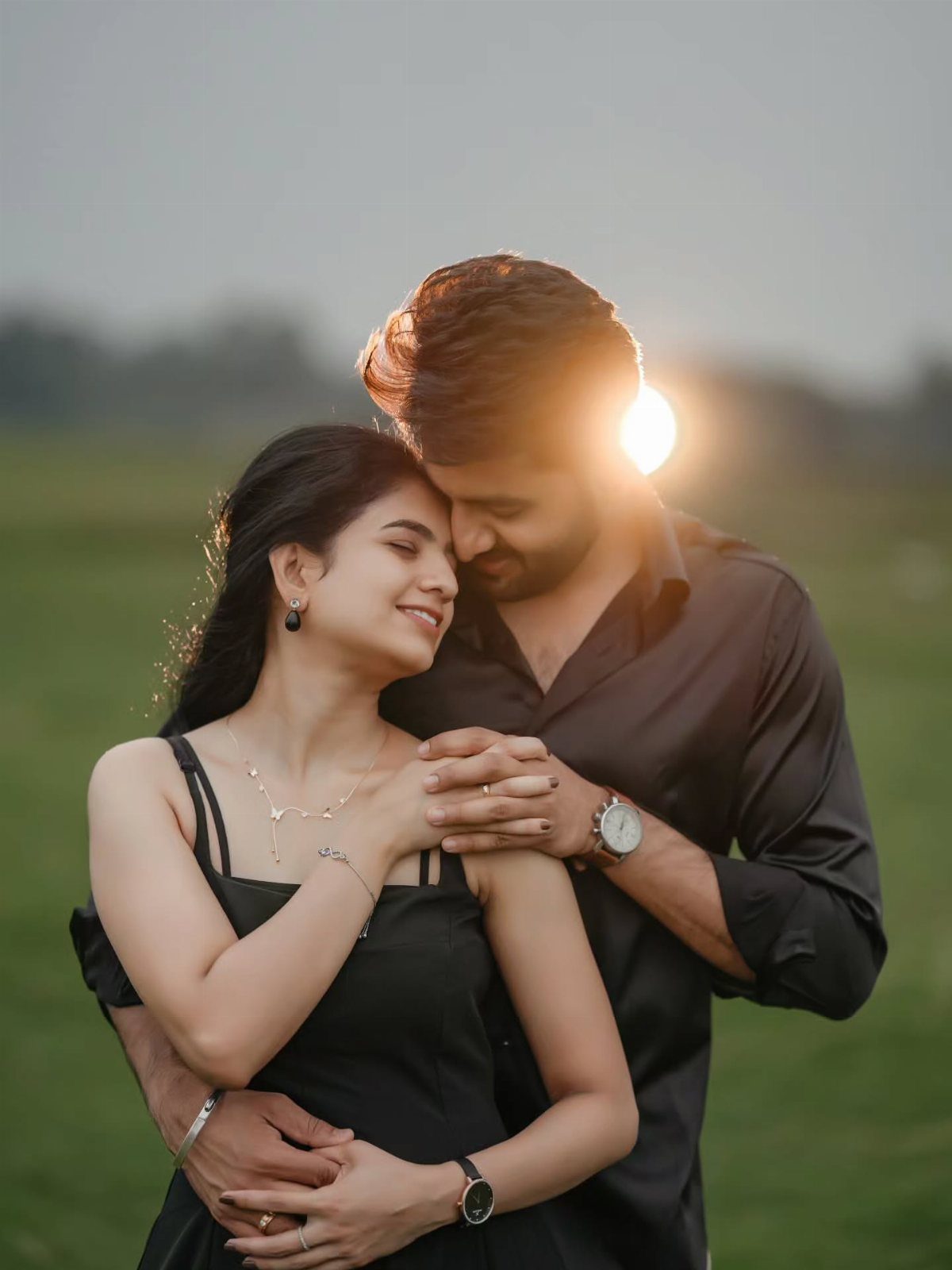 Couple in black clothing embracing in a grassy field during golden hour sunlight.