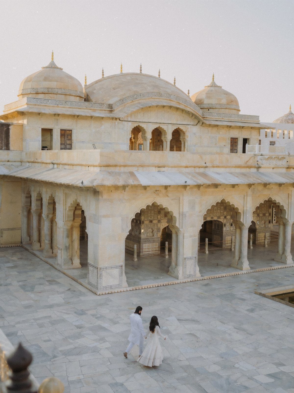 Couple in white traditional clothing walking toward a domed white marble palace courtyard.