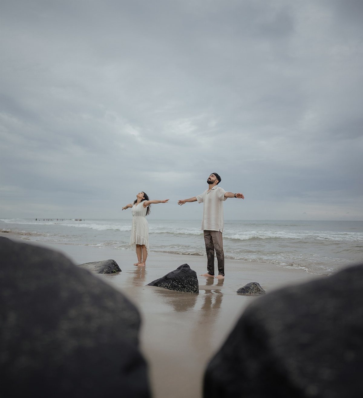 Couple standing on a beach with arms outstretched under a cloudy sky.