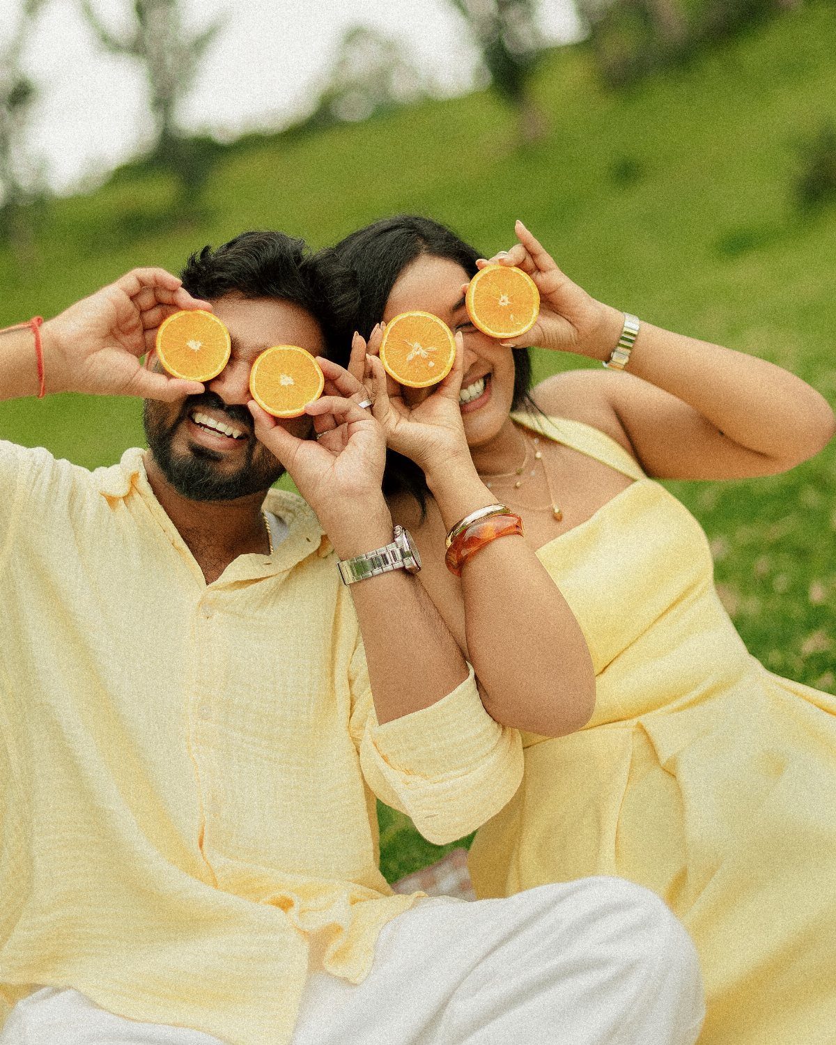 Couple in yellow clothing holding orange slices over their eyes in a grassy field