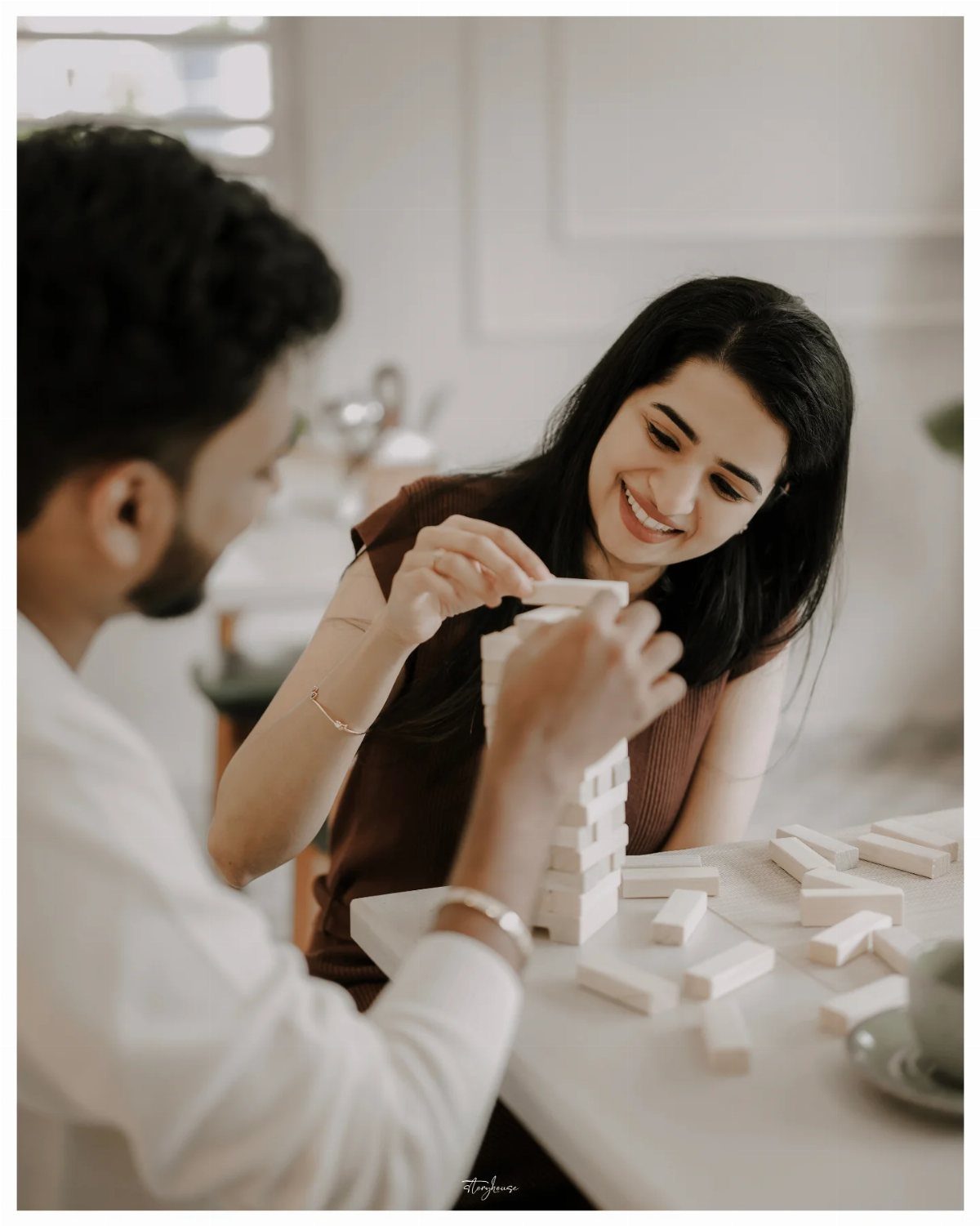 Couple smiling while playing Jenga with wooden blocks on a white table indoors.