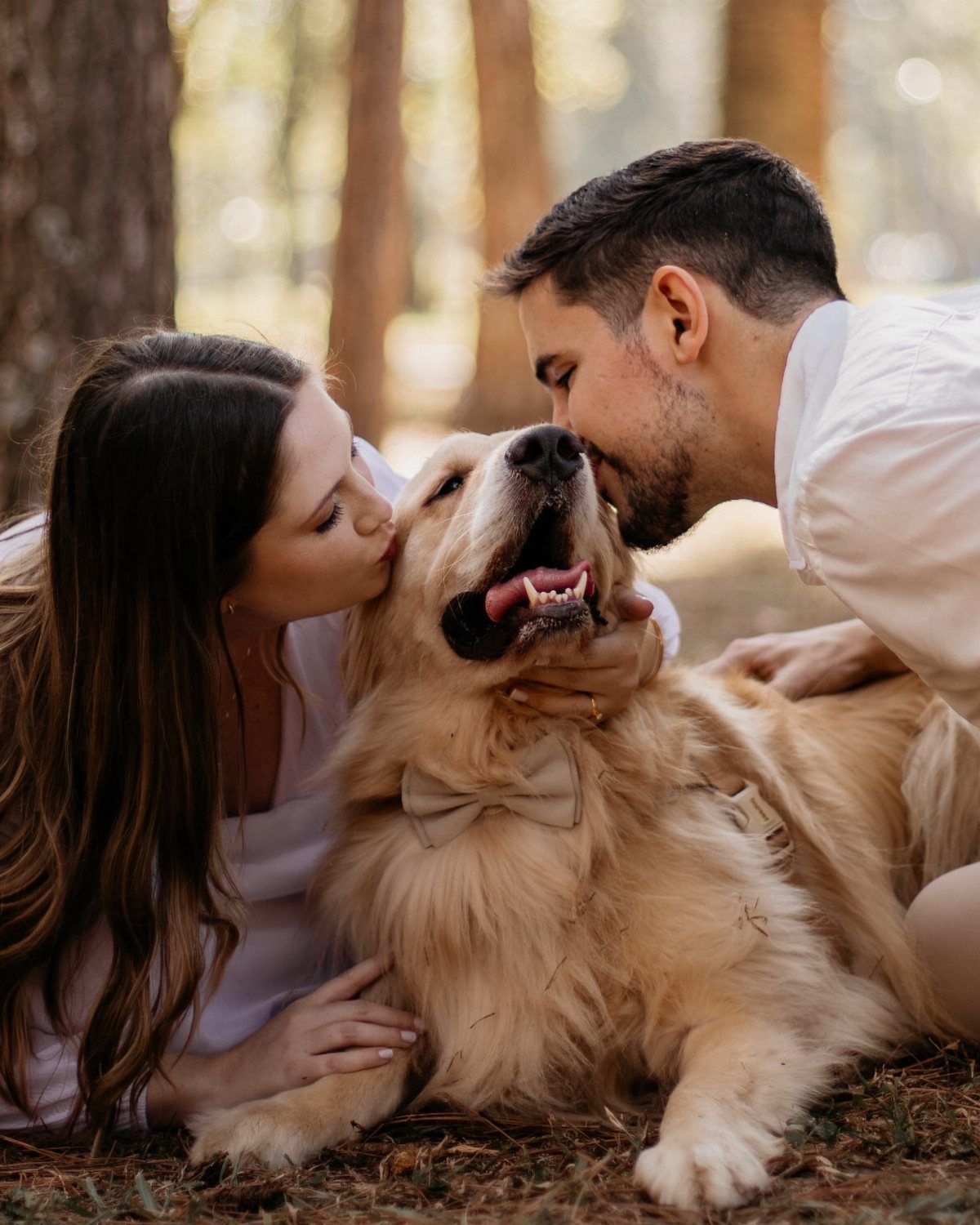 Couple in white kissing their golden retriever wearing a bow tie in the woods
