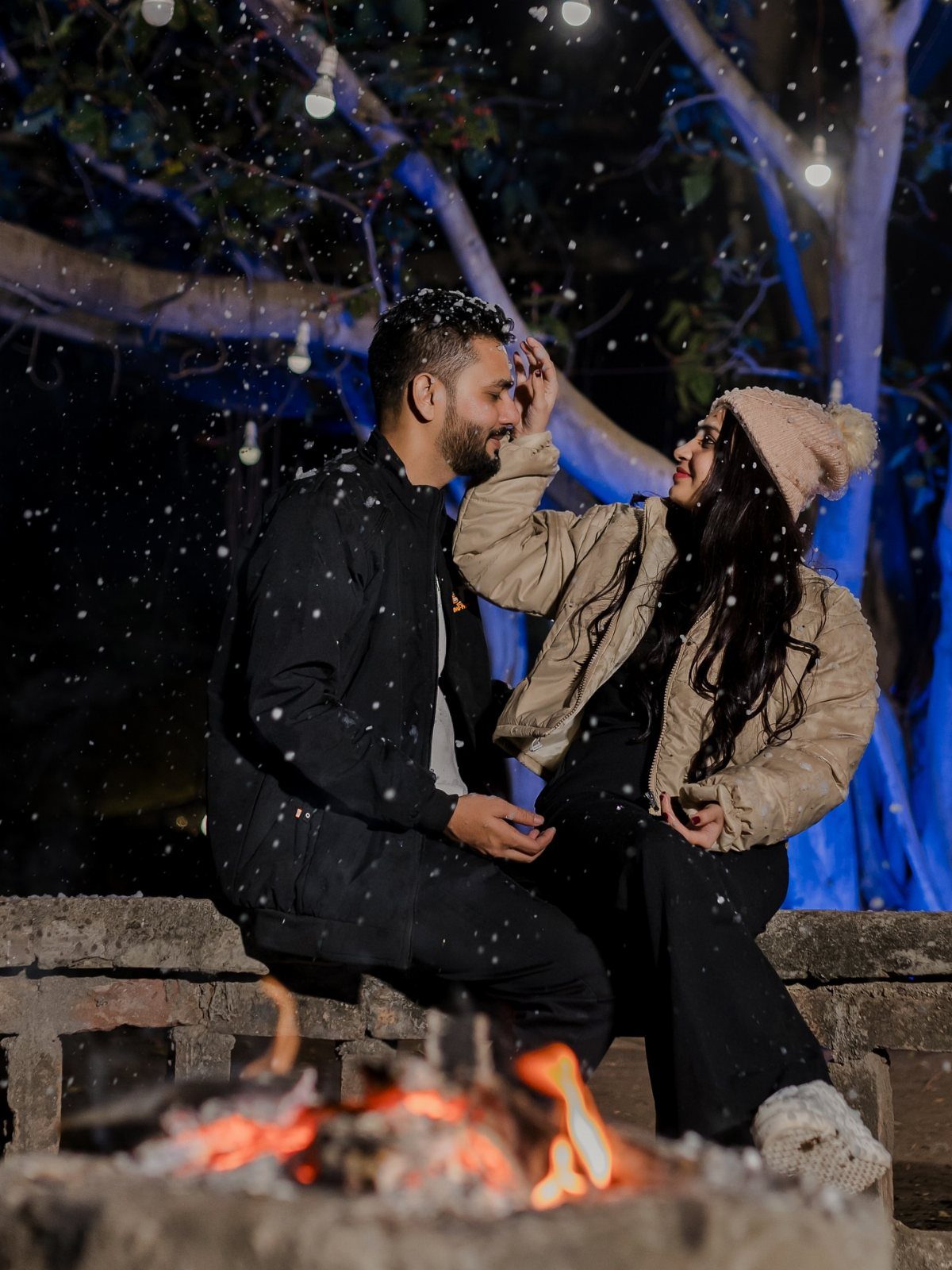 Couple in winter clothing sitting by a campfire with falling snow and blue trees.