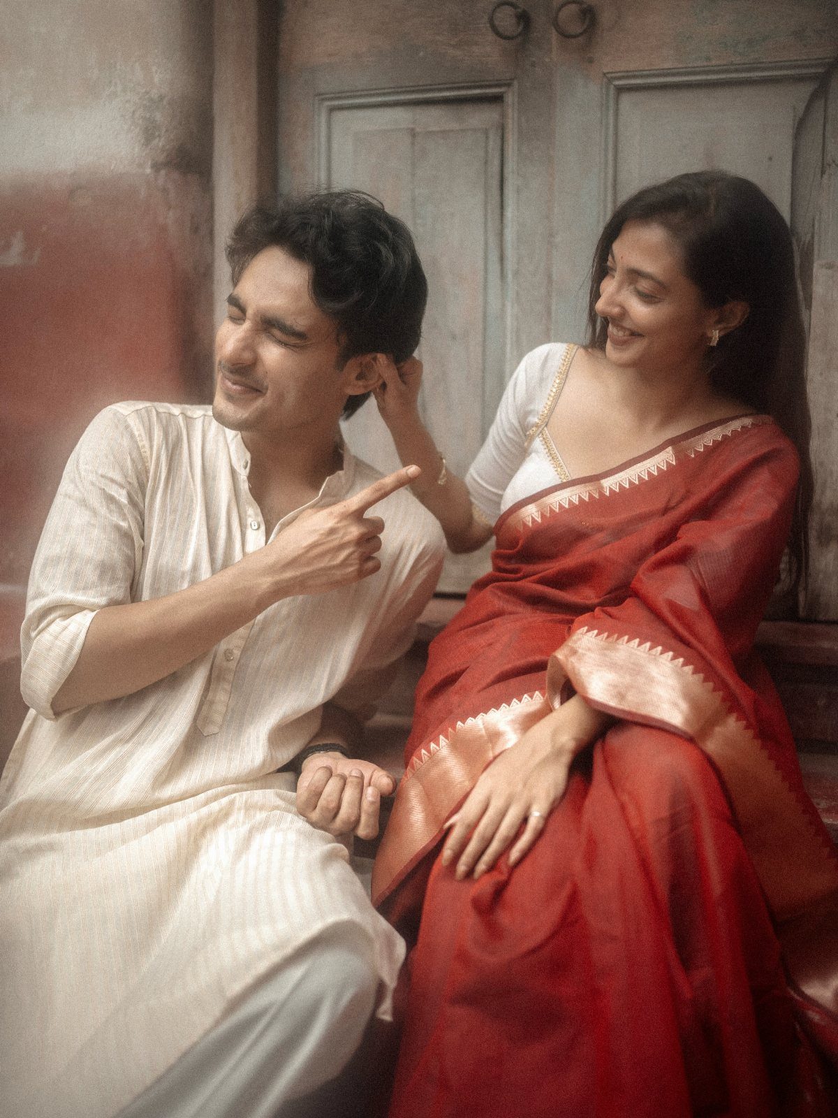Couple in traditional Indian attire posing playfully against a rustic grey wall.