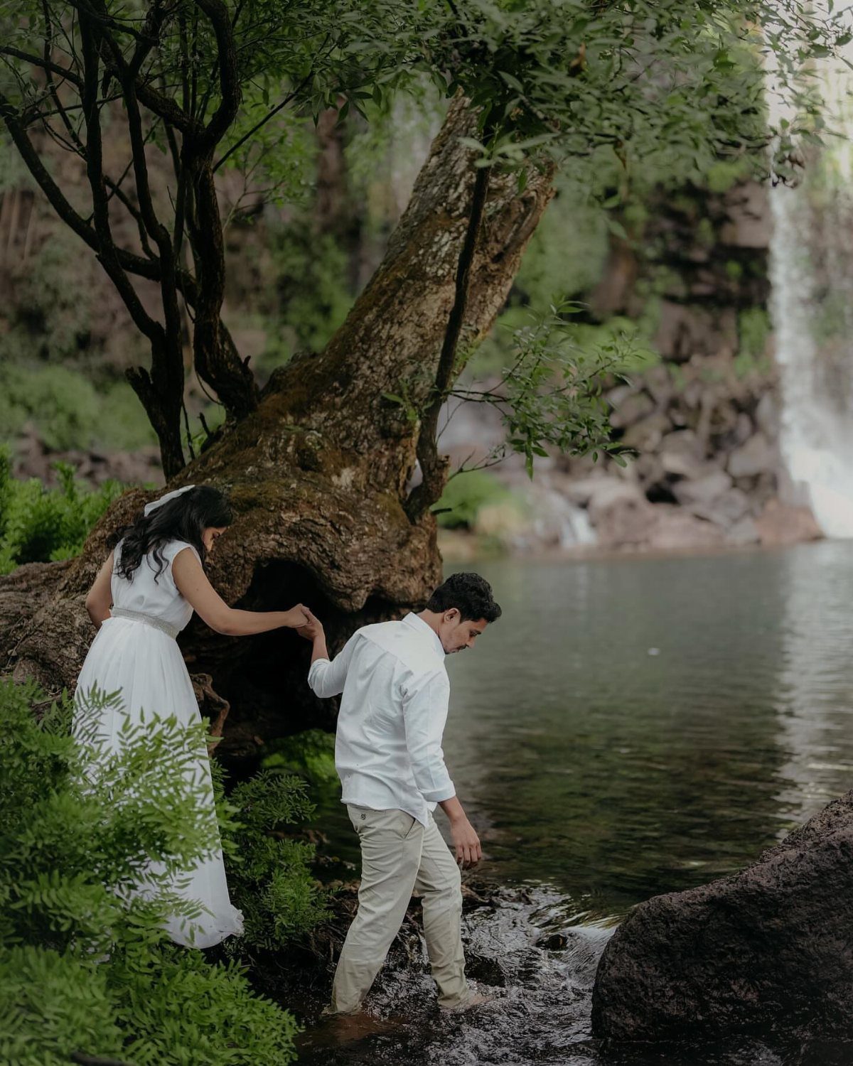 Couple in white clothing by a river with a waterfall and large tree.
