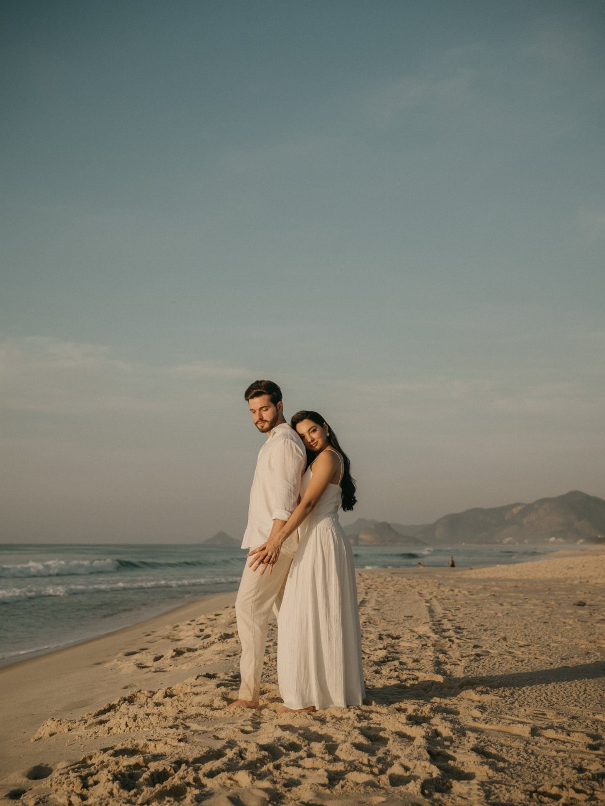 Couple in white outfits embracing on a sandy beach with ocean and mountains.