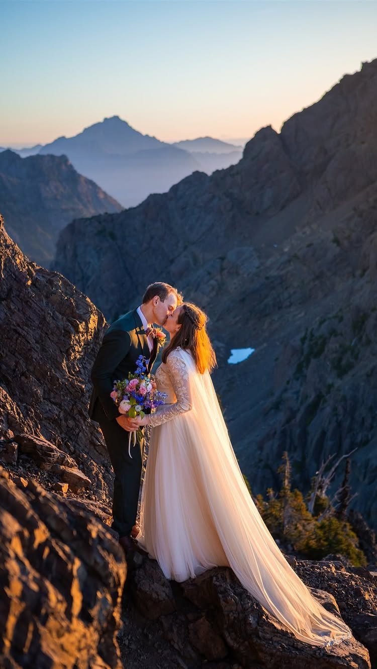 Couple in wedding attire kissing on a rocky mountain peak at sunset