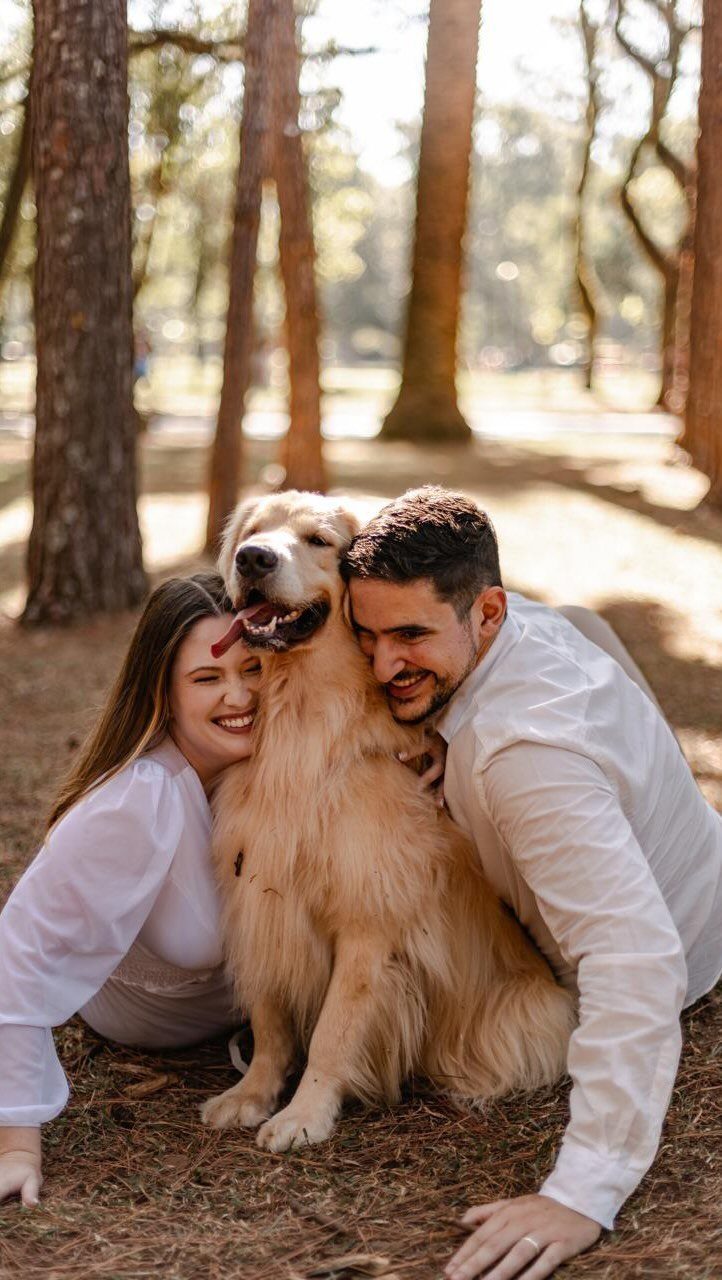 Couple in white outfits hugging a golden retriever in a sunny forest setting