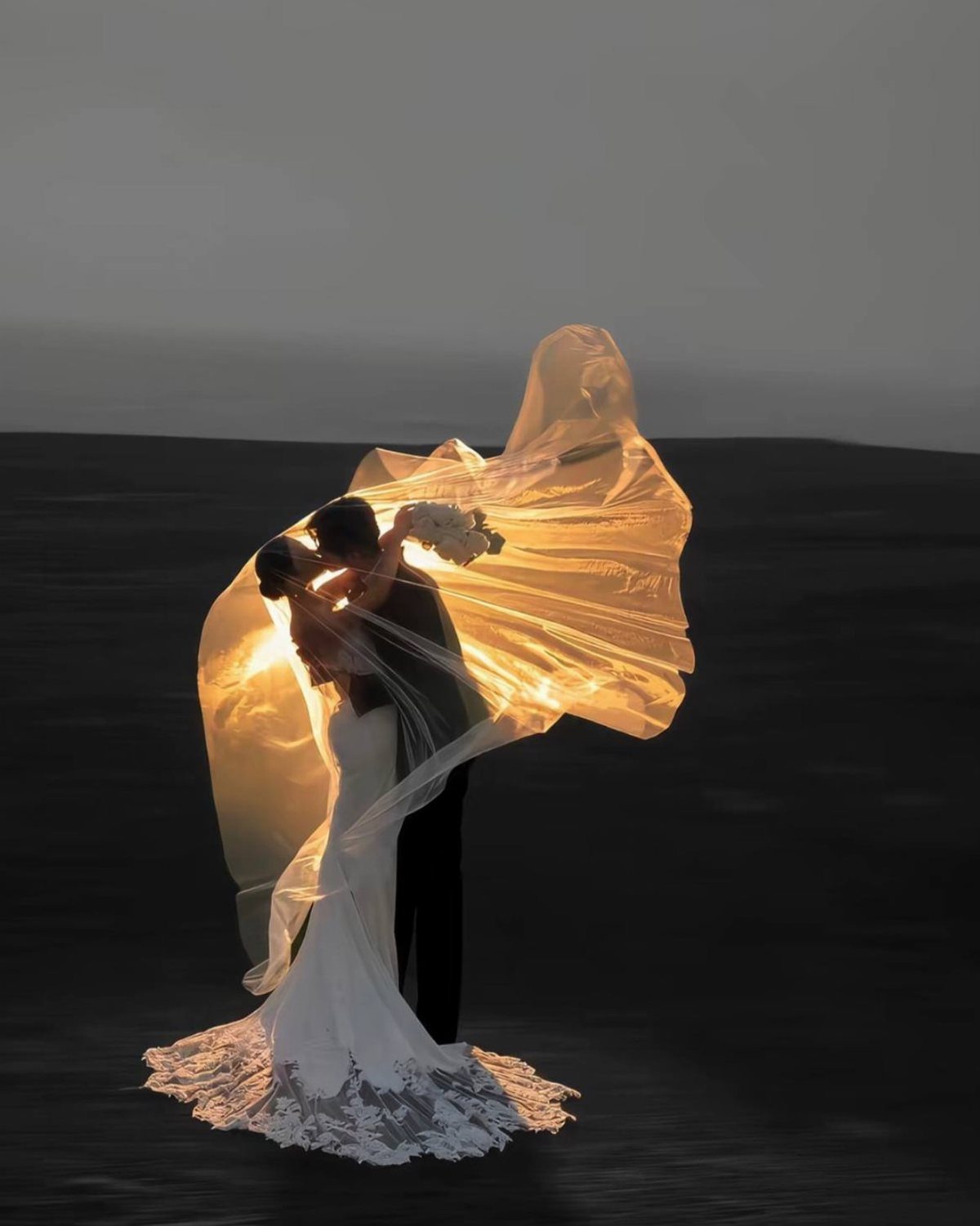 Bride and groom kissing under a glowing, billowing veil in a dark setting