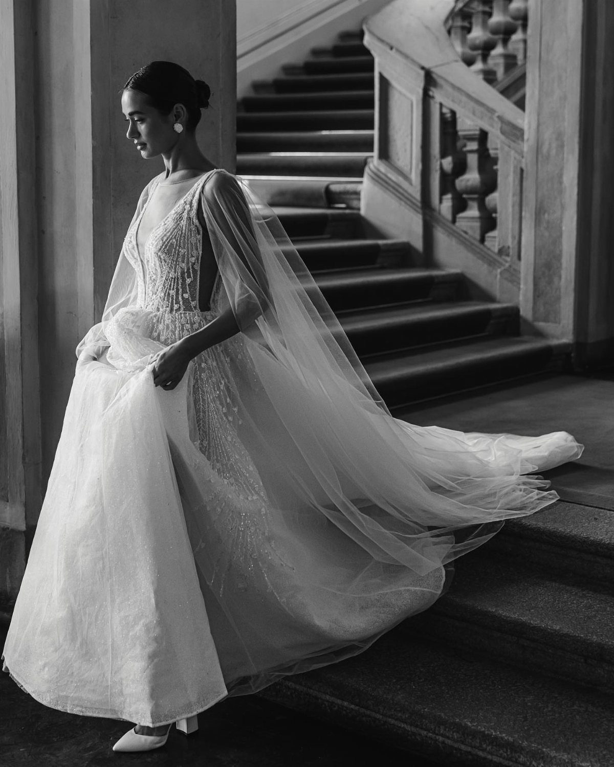 Bride in a white gown and flowing cape standing on stone stairs.