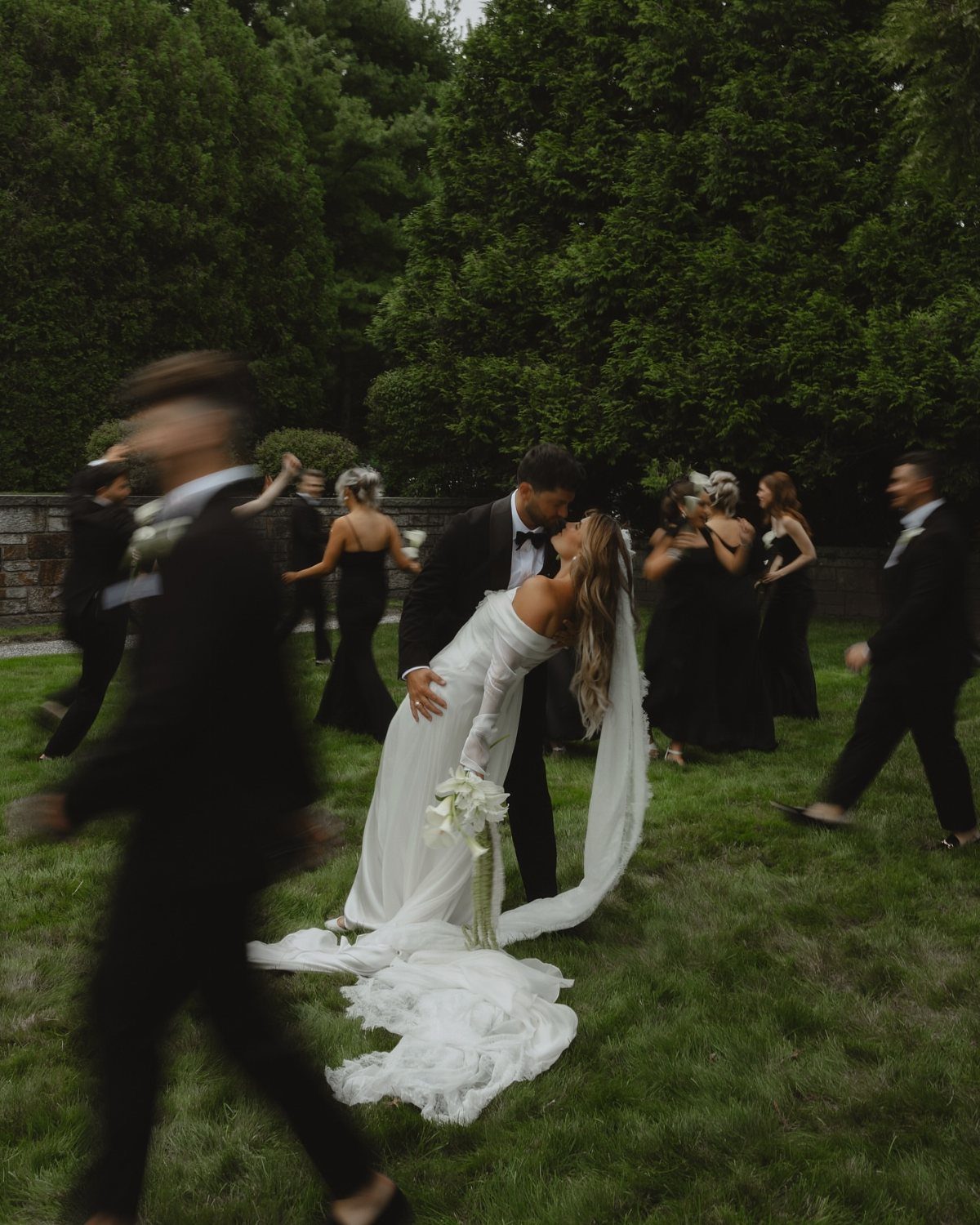 Bride and groom kissing in a garden surrounded by guests in black attire