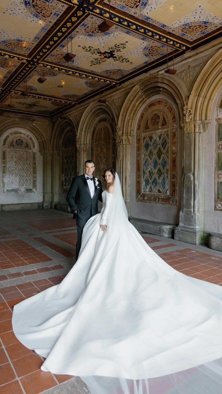 Bride and groom posing in an ornate stone corridor with a decorated ceiling.