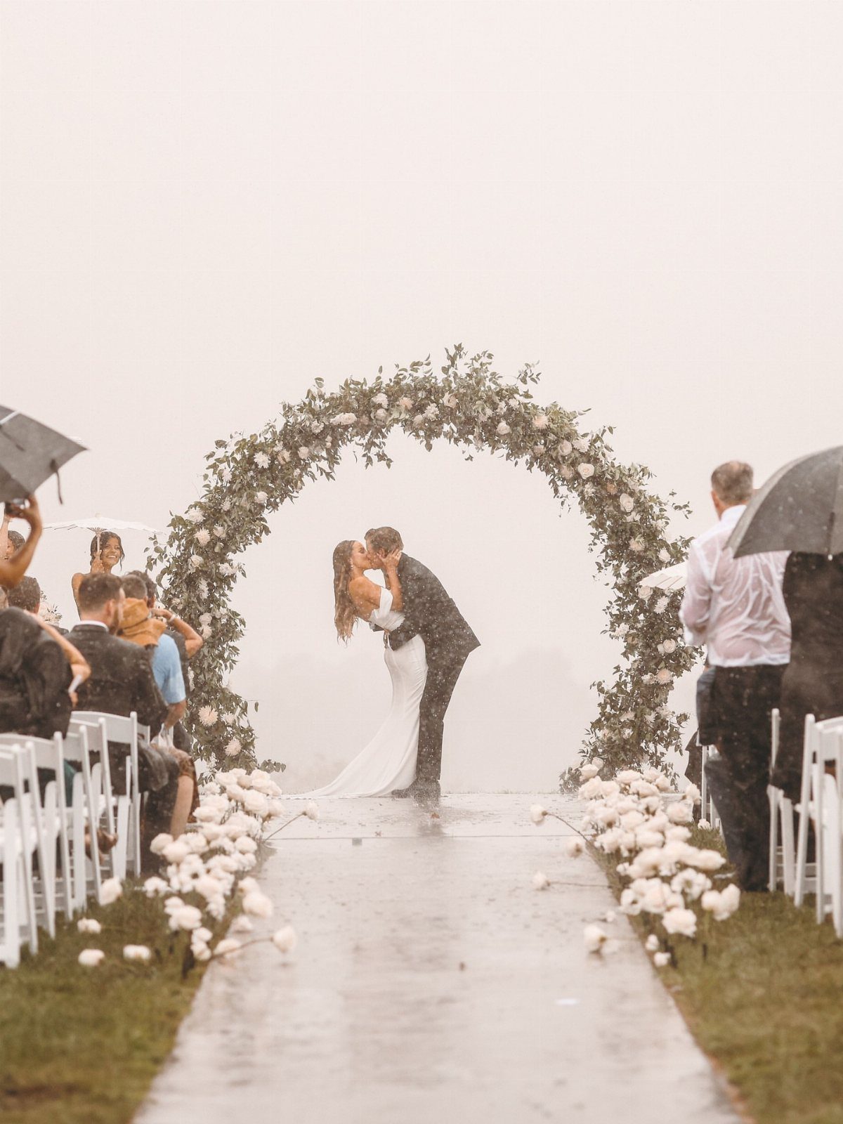 Bride and groom kissing under a floral arch during a rainy outdoor wedding ceremony.
