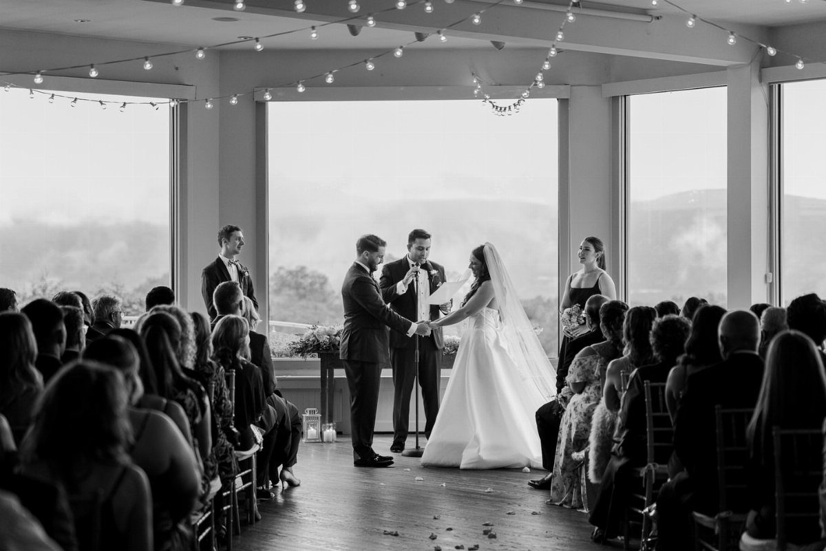 Bride and groom holding hands during an indoor wedding ceremony by large windows.