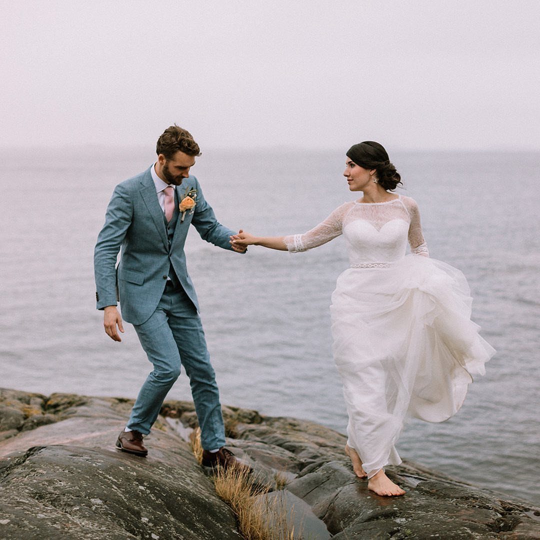 Bride and groom holding hands while walking on wet rocks by the sea.