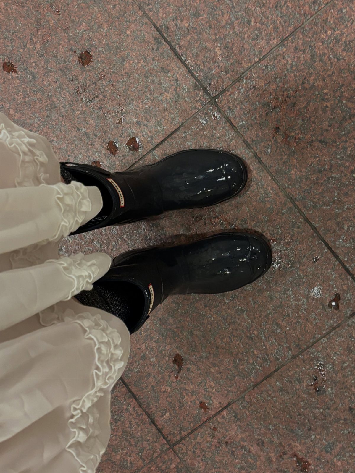 Black glossy rain boots worn under a white ruffled wedding dress on wet tiles.