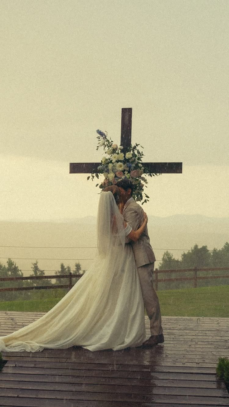 Bride and groom kissing under a floral wooden cross during a rainy wedding.