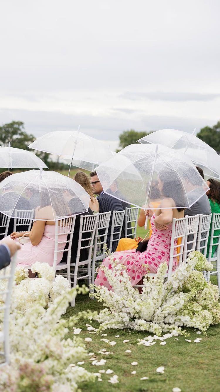 Wedding guests holding clear umbrellas while sitting in white chairs on a lawn.