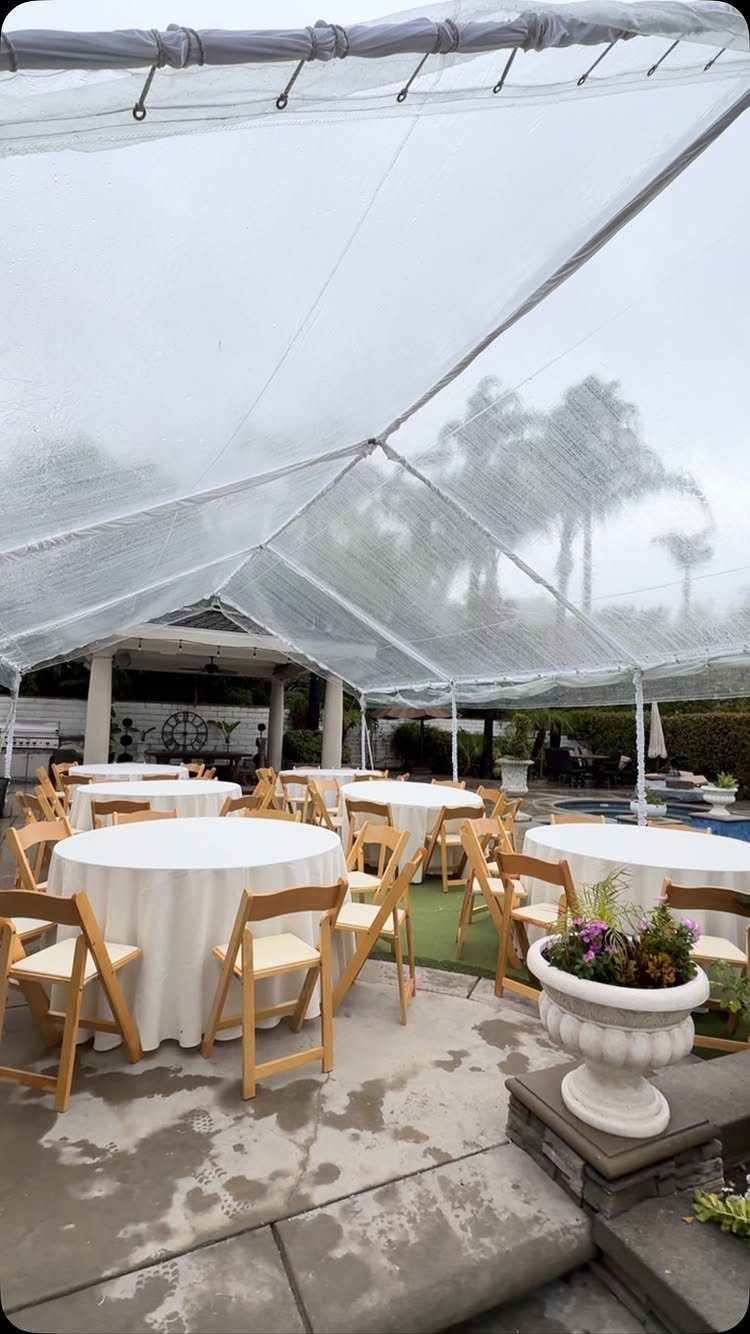 White event tent with clear roof sheltering round tables and chairs on wet concrete.